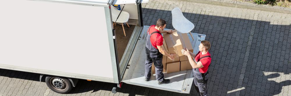 Two workers in red shirts and overalls from Long Island movers are unloading cardboard boxes from a white truck using a hydraulic tail lift. The surrounding area is paved with interlocking stones.